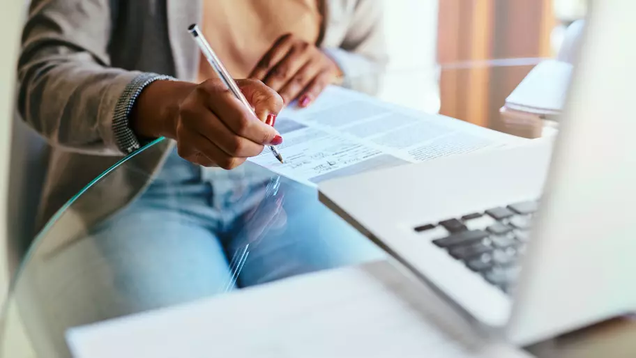 Woman sitting at table filling out a form in front of her laptop.