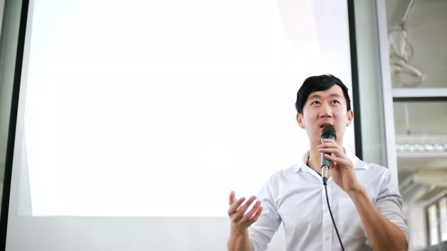 Asian man in white shirt holding a microphone and delivering a speech.