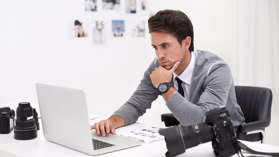 Male photographer sitting at desk with camera and laptop looking somber.