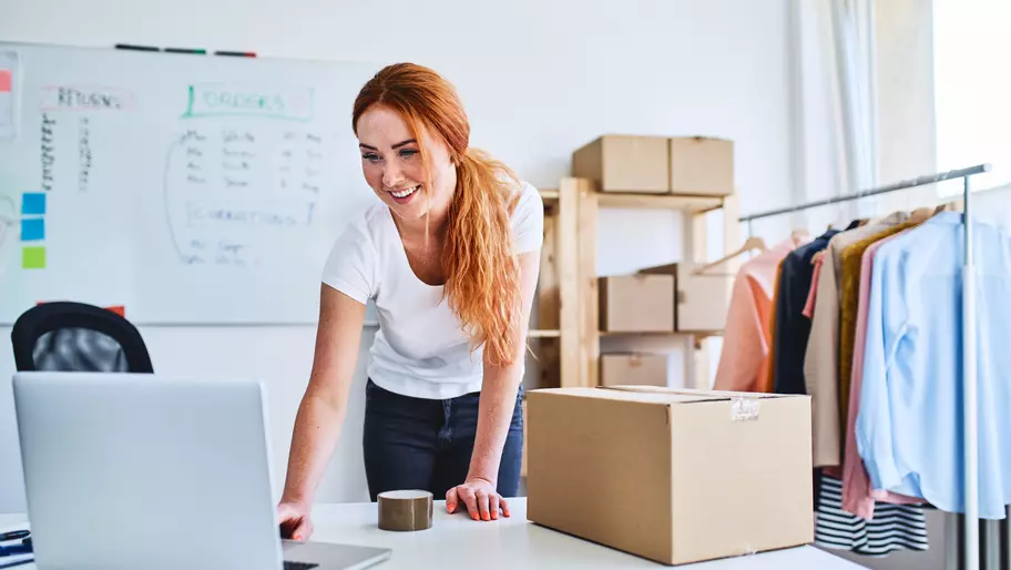 Woman standing over laptop with cardboard box next to her. Evoking the feeling of an entrepreneur or online business owner.