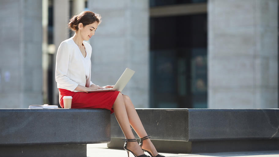 Professional woman in red pencil skirt sitting outside on her laptop.