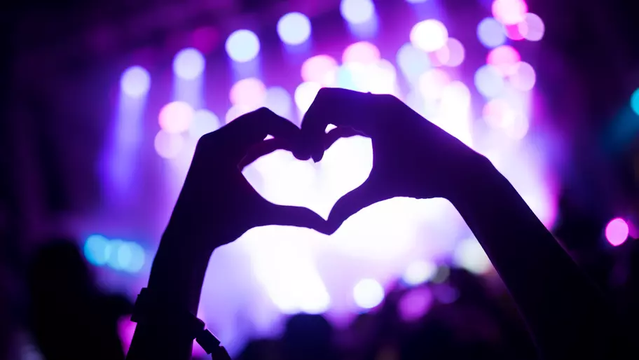 Excited concert crowd with hazy lavender stage in front, one fan makes heart with hands