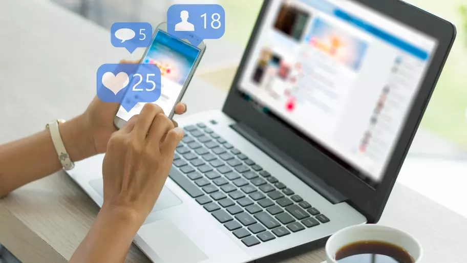 Business owner sitting at laptop using both phone and computer to find relevant Facebook groups