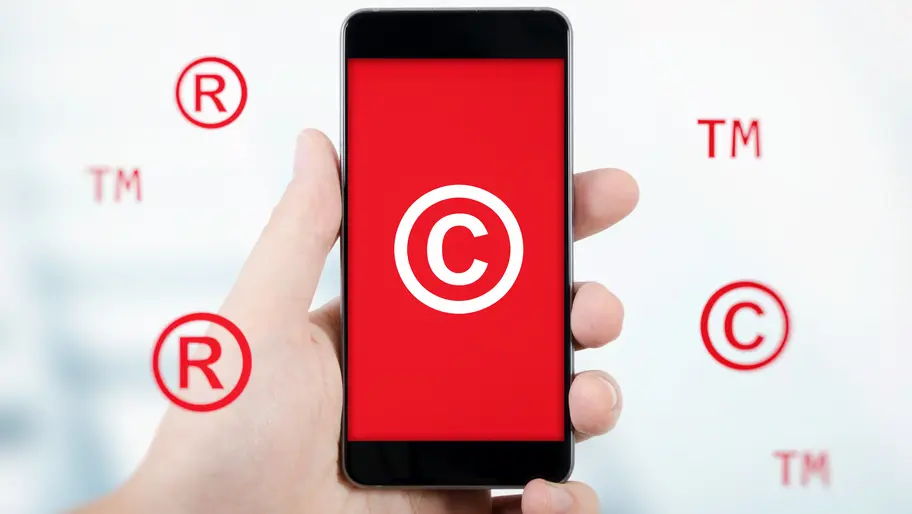 Mobile device in hand displaying red background with trademark and copyright symbols behind the device.
