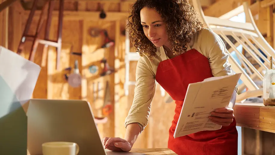 female business owner in red apron leaning over desk looking at SBA changes on her laptop, while holding some business paperwork