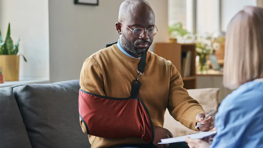 Man sitting on couch wearing tan sweater with his arm in a red sling.