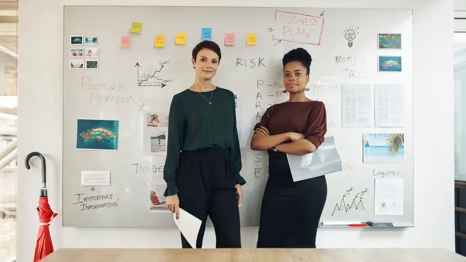 Two buisnesswomen standing in front of a whiteboard filled with strategies and planning