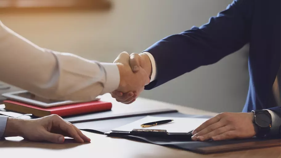 two arms extended in a handshake over a desk with some papers and a red book on it.