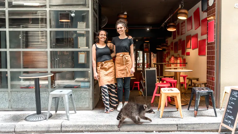 Two café owners pose outside their business as a raccoon walks past the storefront, highlighting an unexpected moment that draws attention.