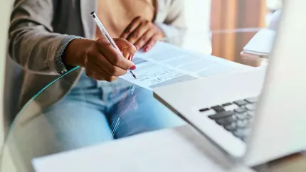 Woman sitting at table filling out a form in front of her laptop.