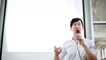 Asian man in white shirt holding a microphone and delivering a speech.