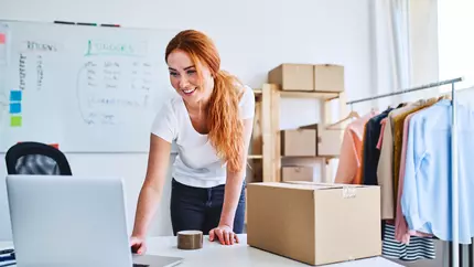 Woman standing over laptop with cardboard box next to her. Evoking the feeling of an entrepreneur or online business owner.