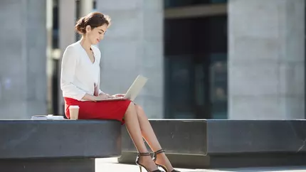 Professional woman in red pencil skirt sitting outside on her laptop.