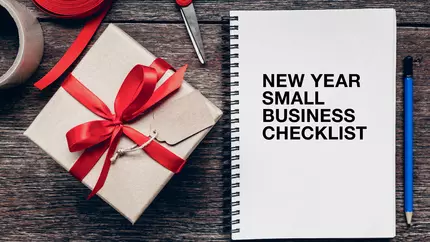 wooden desk with small square gift with red ribbon sitting next to spiral notebook with new year small business cheklist printed on the cover
