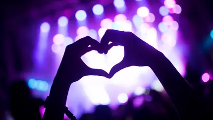 Excited concert crowd with hazy lavender stage in front, one fan makes heart with hands
