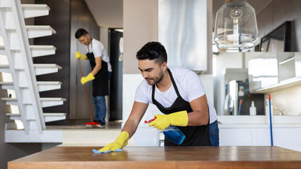 two men with yellow cleaning gloves sweeping floor and dusting table in a bright white room