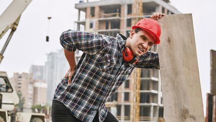 male small business construction worker bending slightly holding back with building in background.