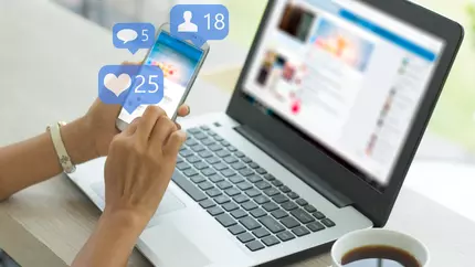 Business owner sitting at laptop using both phone and computer to find relevant Facebook groups