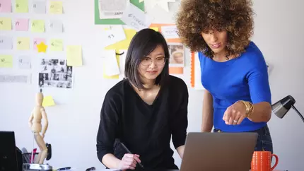female business owners at a desk with whiteboard behind them, mentoring one another on best practices