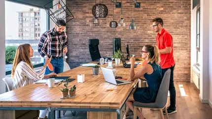 A stress-free business enviroment with two female employees sitting and two male employees playing ping pong while discussing buisness matters at conference table
