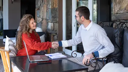 Female in red shirt shaking hands with male in grey shirt at a casual business meeting outdoors.
