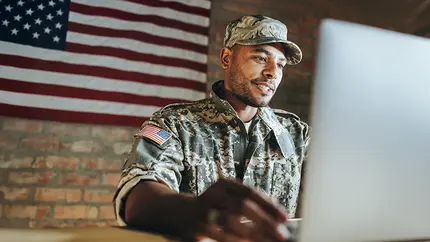 US veteran small business owner sitting at laptop with American flag behind him, registering his VOSB