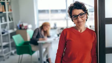 Business woman with dark hair and glasses in red blouse standing at the front right hand side with colleagues in the background on the left