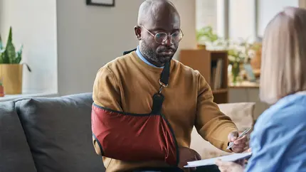 Man sitting on couch wearing tan sweater with his arm in a red sling.