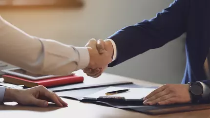 two arms extended in a handshake over a desk with some papers and a red book on it.