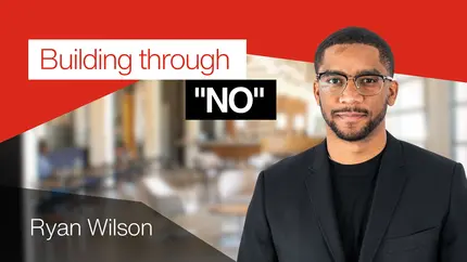 Graphic featuring the title “Building through ‘NO’” in bold text on a red and white background, with Ryan Wilson’s name displayed in the lower-left corner. The background shows a blurred office setting.