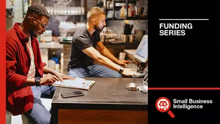 two men filling out grant applications in their small business location - one using paper and pen, the other on a computer.