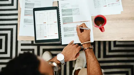 Top-down view of a small business owner reviewing budgeting documents and financial spreadsheets on a desk using a tablet and pen.