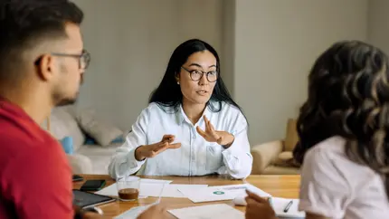Life coach leading a coaching session with two clients at a table, discussing plans and reviewing documents during a one-on-one meeting.