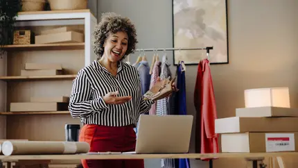 Female small‑business owner presenting product samples during an online meeting in her boutique workspace, surrounded by packaging materials and clothing racks, illustrating how proactive protection supports sustainable business growth.