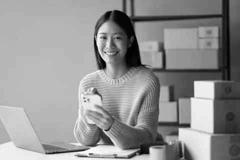 An online retailer taps on phone and sits at desk with shipping boxes