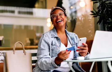 Smiling person sits at café table holding phone and credit card by laptop