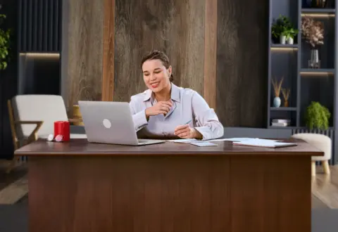 Woman smiling at laptop, taking notes at desk