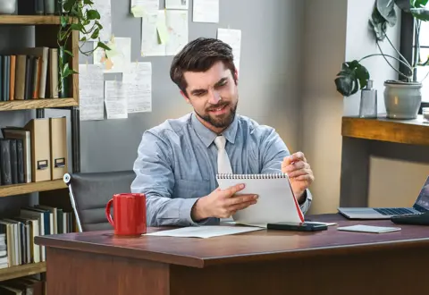 Man writing in a notebook at an office desk with coffee mug